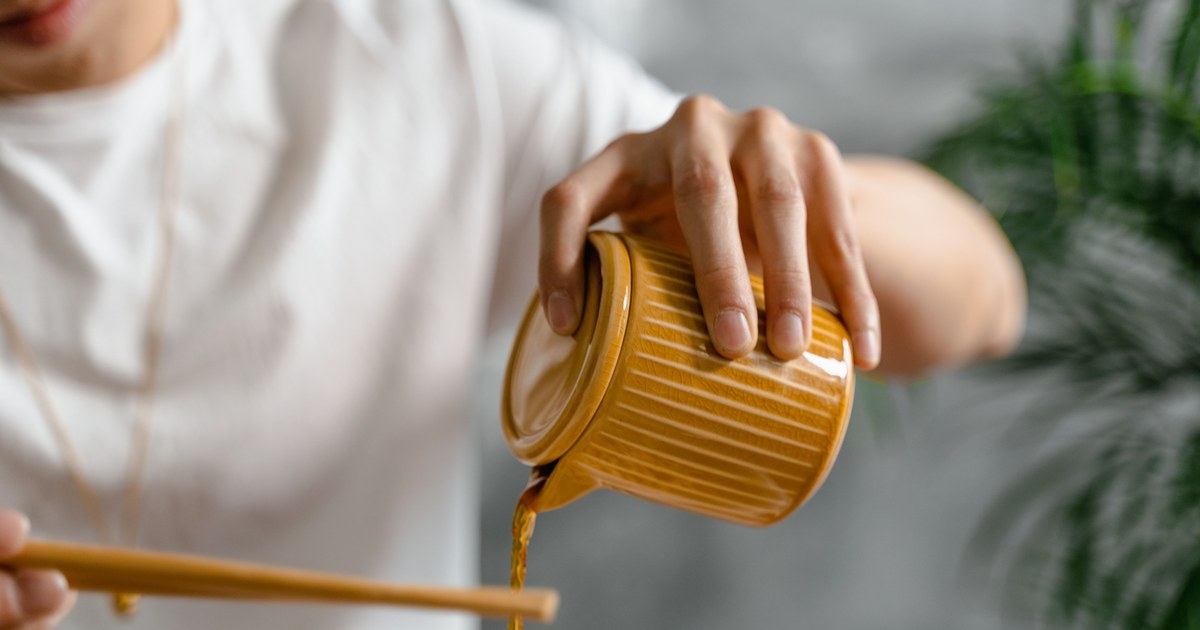 A hand pouring soy sauce from a ceramic jug over a bowl of noodles